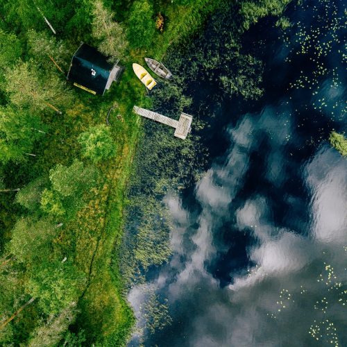 Aerial view of sauna house by the lake shore. Wooden pier with fishing boats.